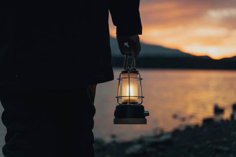 a person holding a lantern near a body of water, warmly lit, warm coloured, explorer, lantern candle