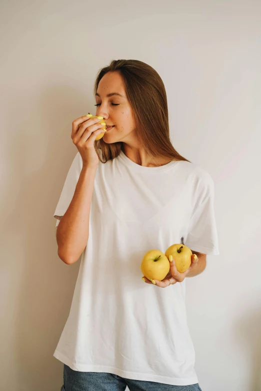 a woman standing in front of a white wall eating an apple, by Nicolette Macnamara, trending on unsplash, wearing a modern yellow tshirt, wearing white pajamas, organics, manuka