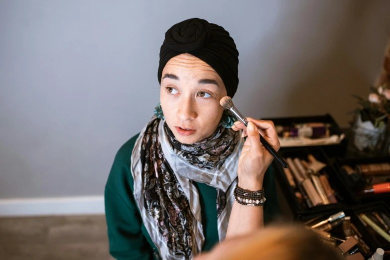 a woman is doing makeup in front of a mirror, by Julia Pishtar, hurufiyya, wearing a head scarf, androgynous male, mid shot photo, commercially ready
