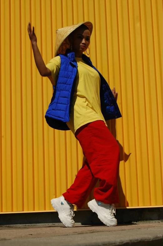 a woman walking down a sidewalk in front of a yellow wall, red tank top and wide blue pants, hip hop style, federation clothing, in an action pose