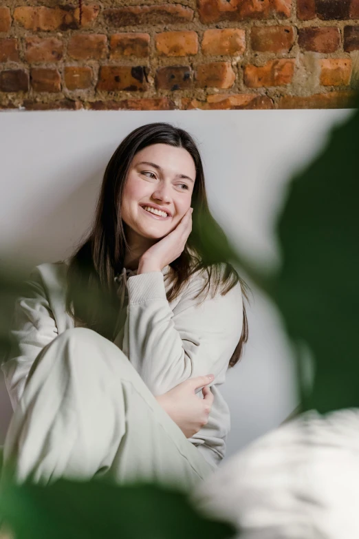 a woman sitting on top of a bed next to a plant, trending on pexels, soft smile, wearing white pajamas, portait image, happy grin