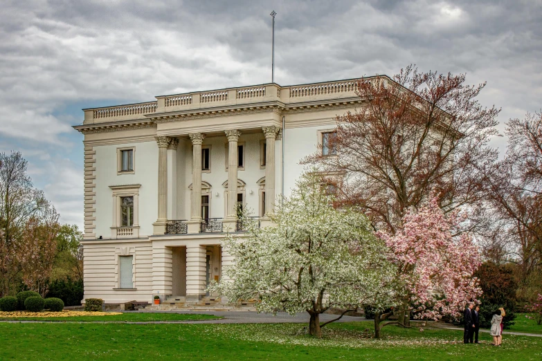 a large white building sitting on top of a lush green field, by Harry Haenigsen, neoclassicism, sakura, us journalism ministry photo, on a cloudy day, decorated with soviet motifs