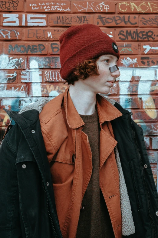 a man standing in front of a graffiti covered wall, an album cover, trending on pexels, ginger hair with freckles, maroon hat, model wears a puffer jacket, very very very pale skin