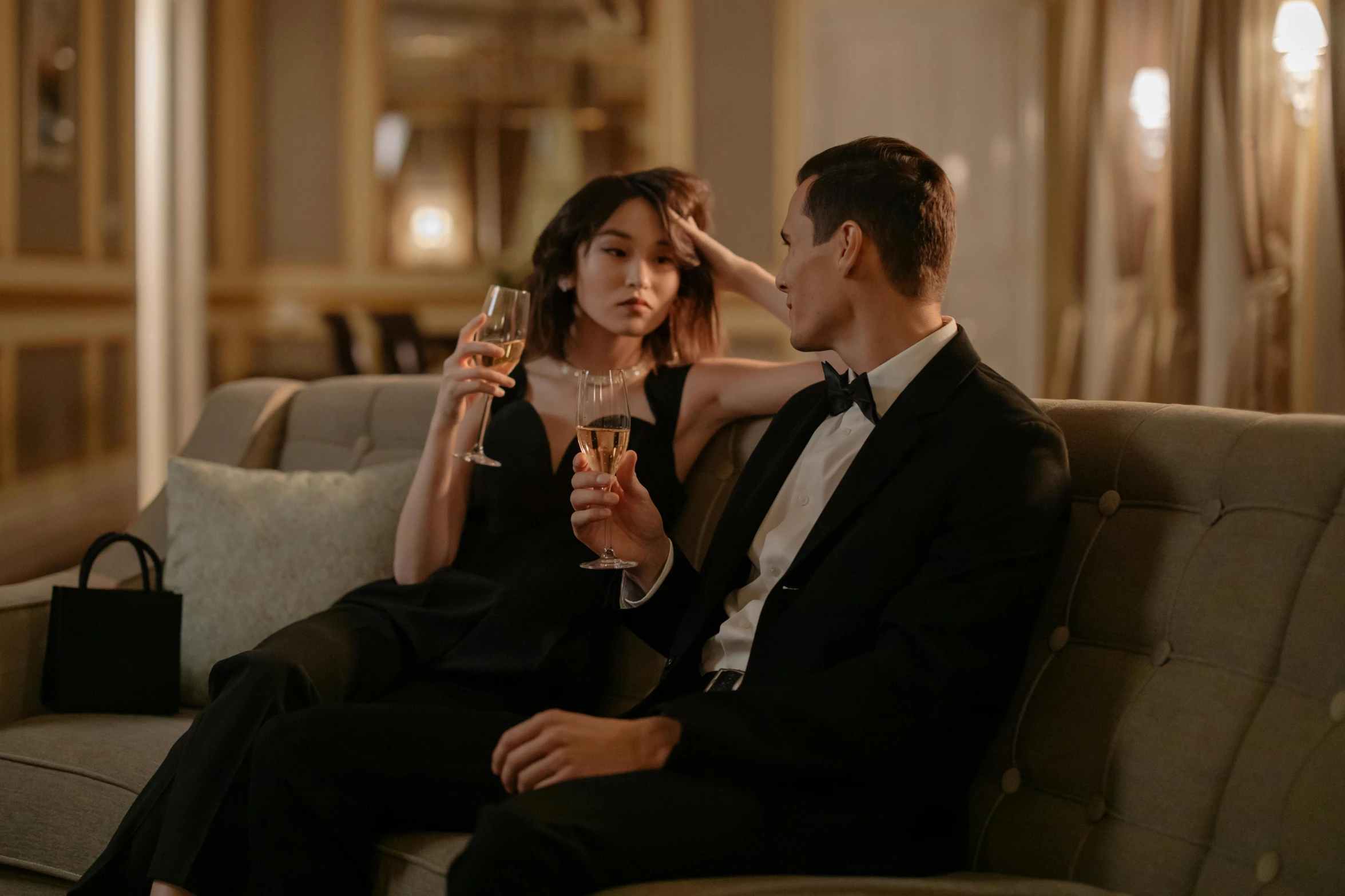 a man and a woman sitting on a couch holding wine glasses, gemma chan, tailored clothing, private moment, hotel room