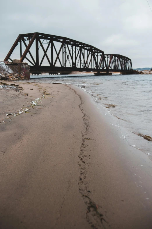 a train bridge over a body of water, footprints in the sand, superior, ((rust)), windy beach