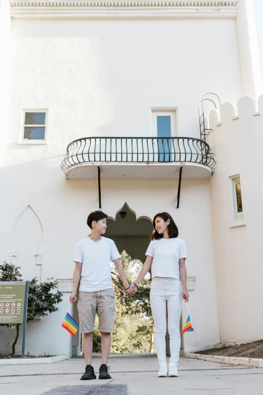 a man and woman holding hands in front of a building, a colorized photo, by Okuda Gensō, unsplash, palm springs, dressed in a white t-shirt, in a castle, pride month