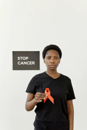 a woman holding a red ribbon in front of a stop cancer sign, a photo, by Lily Delissa Joseph, pexels contest winner, renaissance, dark skinned, courtesy of moma, on a gray background, wearing an orange t-shirt