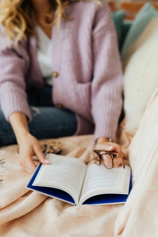 a woman sitting on a bed reading a book, by Nicolette Macnamara, trending on pexels, wearing square glasses, warmer and softer colours, indigo, seasonal