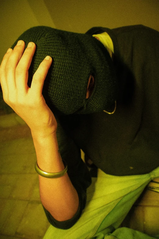 a person sitting on a toilet wearing a hat, hands shielding face, green dress with a black hood, ((oversaturated)), in a dark green polo shirt