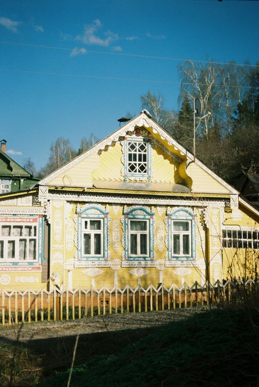 a yellow house with a fence in front of it, inspired by Konstantin Vasilyev, art nouveau, log houses built on hills, decorated with russian motifs, photo taken on fujifilm superia, square