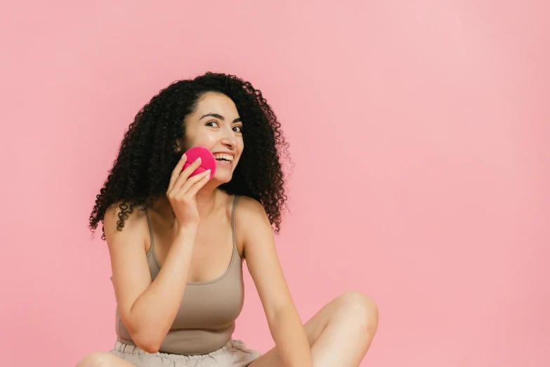 a woman sitting on the floor eating an apple, trending on pexels, figuration libre, pink background, girl making a phone call, with textured hair and skin, the woman holds more toys