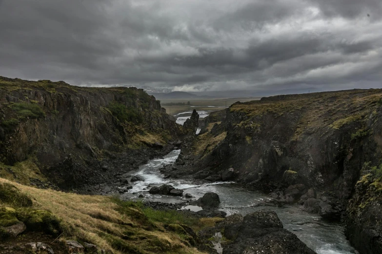 a man standing on top of a cliff next to a river, by Þórarinn B. Þorláksson, pexels contest winner, hurufiyya, grey cloudy skies, ravine, bridge, panoramic shot