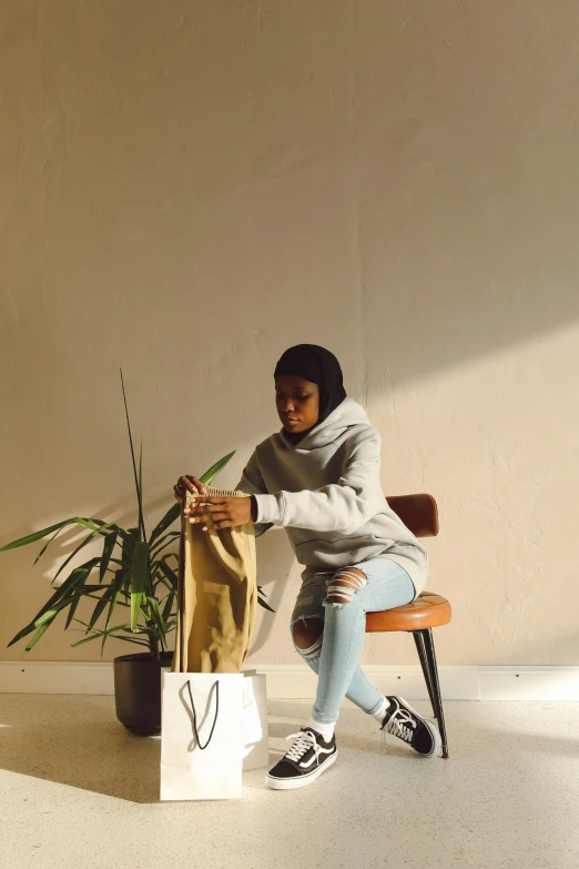 a woman sitting on a bench next to a plant, by Lily Delissa Joseph, beige hoodie, exiting store, in a studio, ameera al-taweel