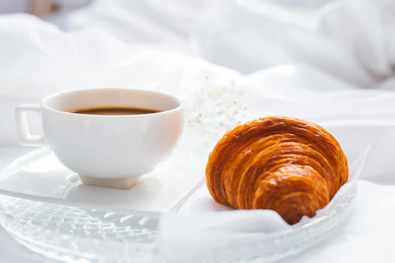 a white plate topped with a croissant next to a cup of coffee, inspired by Charles Le Roux, trending on unsplash, romanticism, white sheets, twinkling and spiral nubela, background image