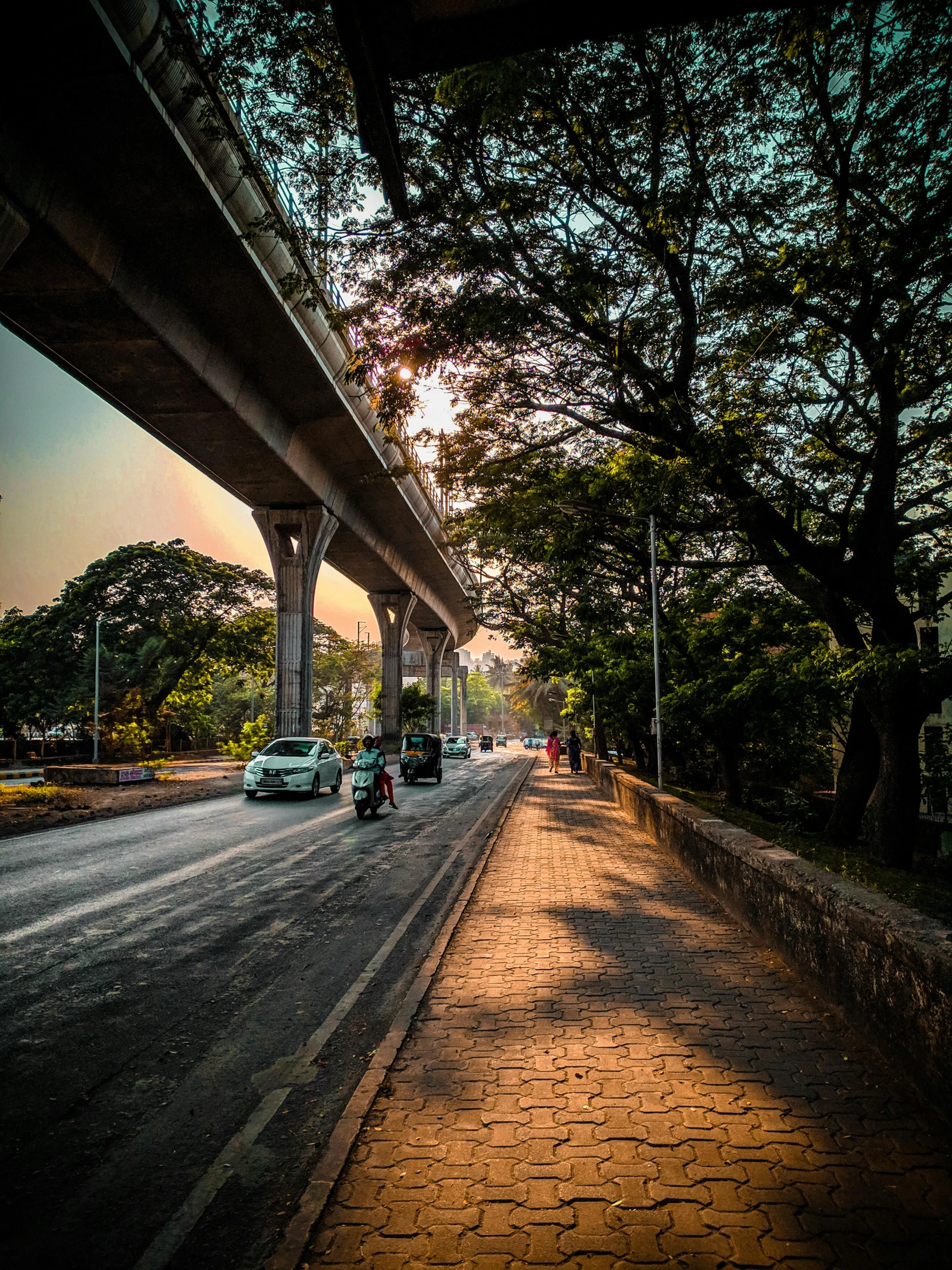 a group of cars driving down a street next to a bridge, by Max Dauthendey, pexels contest winner, guwahati, tree-lined path at sunset, sitting under bridge, bangalore