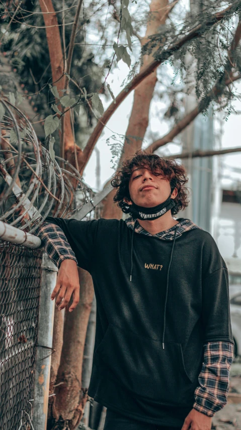 a man standing next to a chain link fence, an album cover, by Everett Warner, trending on pexels, graffiti, wavy hair spread out, hoodie, amongst foliage, teen boy