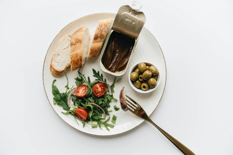 a white plate topped with a sandwich and a bowl of olives, by Nicolette Macnamara, pexels contest winner, art nouveau, sardine in a can, clean minimalist design, metallic, salad