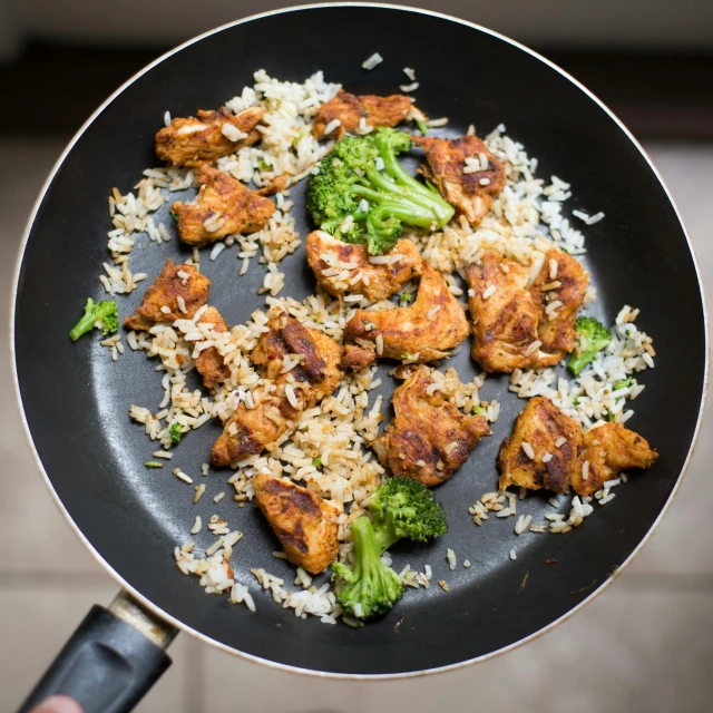 a person holding a frying pan filled with rice and broccoli, chicken, image, satisfying, textural