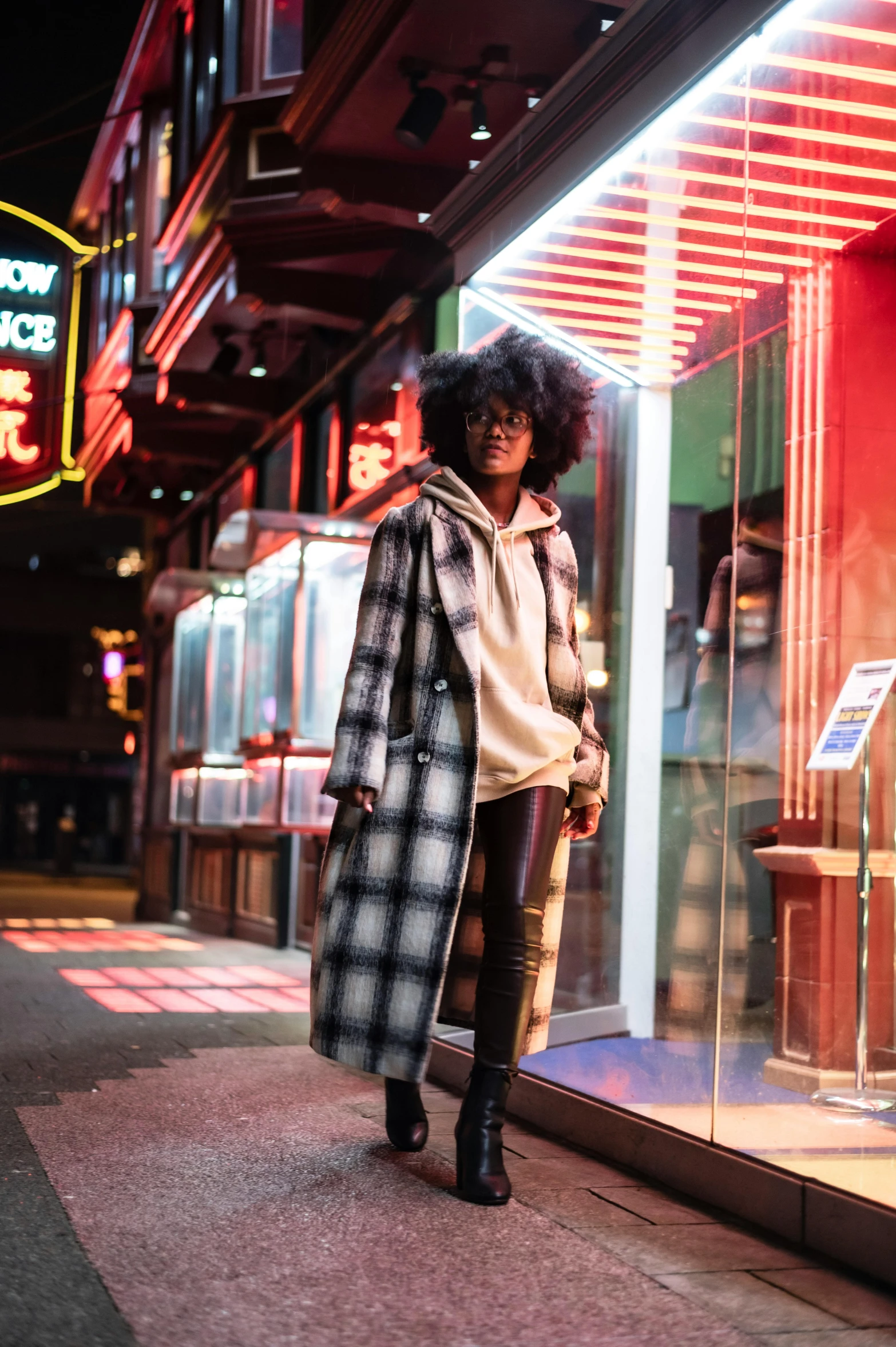 a woman standing in front of a store window, trending on unsplash, realism, tartan hoodie, harlem, lights, long coat