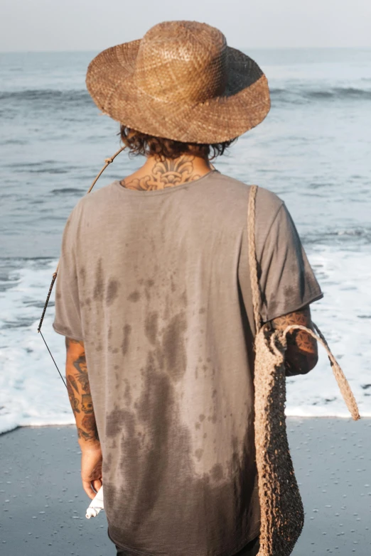 a man standing on top of a beach next to the ocean, dirty short sleeved shirt, faded hat, profile image, tattooed back