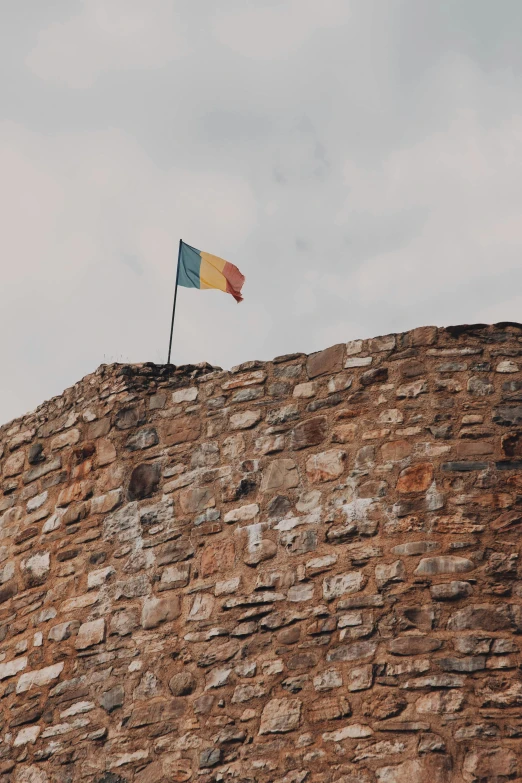 a stone wall with a flag on top of it, a photo, by Matija Jama, trending on unsplash, renaissance, romanian heritage, multiple stories, ancient ruins behind her, brussels