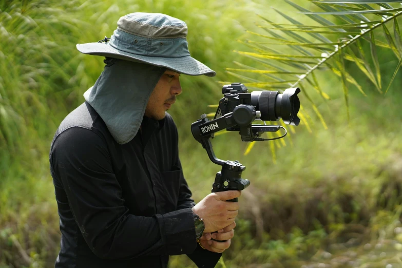 a man in a hat holding a camera on a tripod, a picture, inspired by Kanō Tan'yū, unsplash, sumatraism, still from nature documentary, shot with sony alpha 1 camera, youtube thumbnail, sony a 7 siii