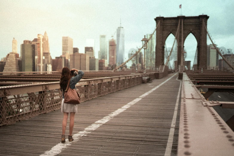 a woman walking across a bridge with buildings in the background, inspired by Michael Komarck, pexels contest winner, photorealism, watching new york, vintage color photo, panoramic view of girl, medium format