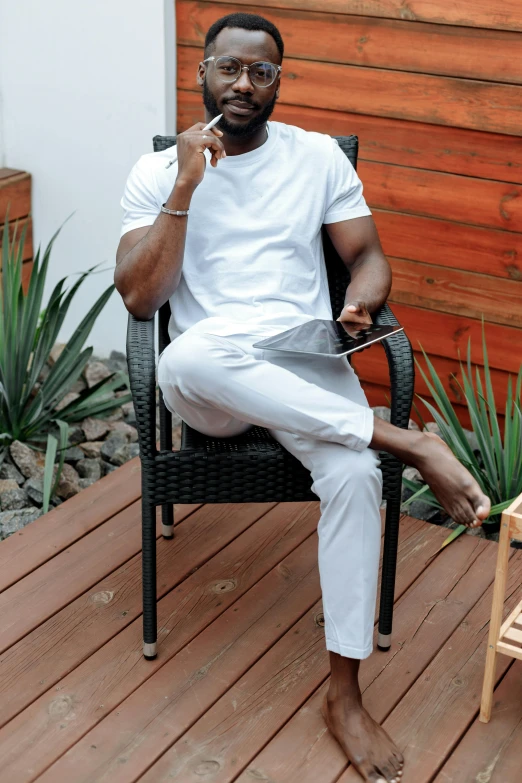 a man sitting in a chair smoking a cigarette, white pants, plain white tshirt, adebanji alade, next to a plant