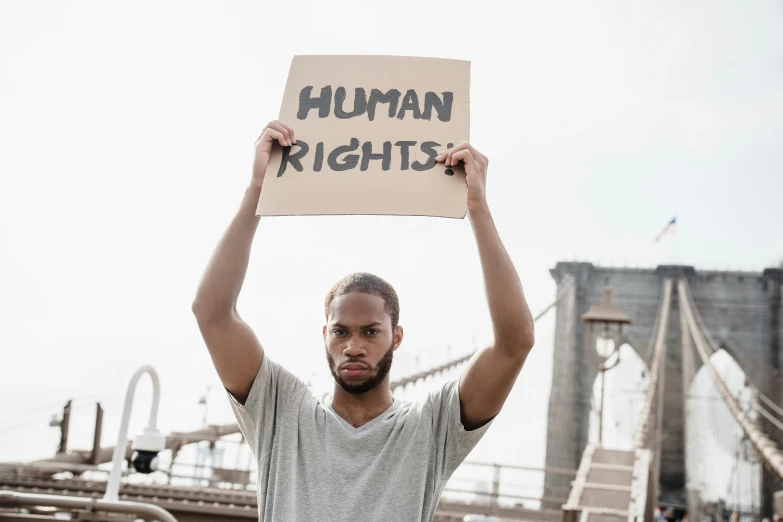 a man holding a sign that says human rights, by Julia Pishtar, trending on pexels, black arts movement, high bridges, brown, profile picture, light skin