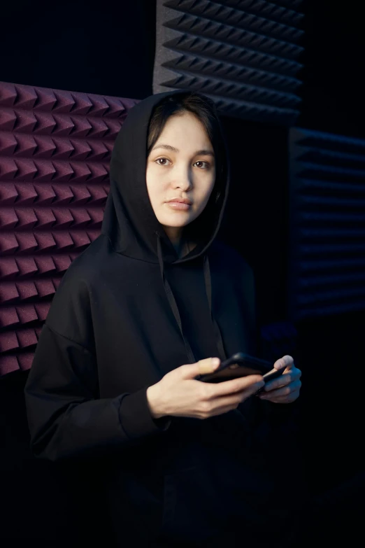 a woman in a black hoodie holding a cell phone, in a studio, vantablack cloth technology, kiko mizuhara, ameera al taweel