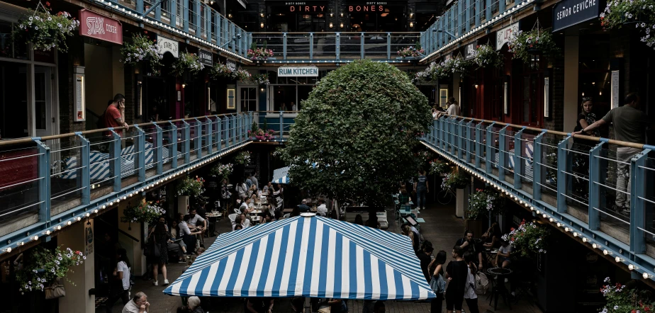 a blue and white striped umbrella sitting inside of a building, inspired by Thomas Struth, unsplash, people sitting at tables, central tree, marketsquare, te pae