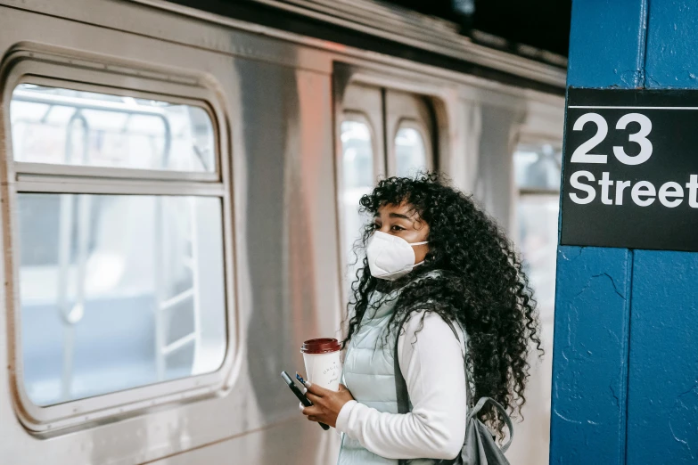 a woman with a face mask standing next to a train, by Carey Morris, trending on pexels, in new york city, avatar image, coffee smell, sza