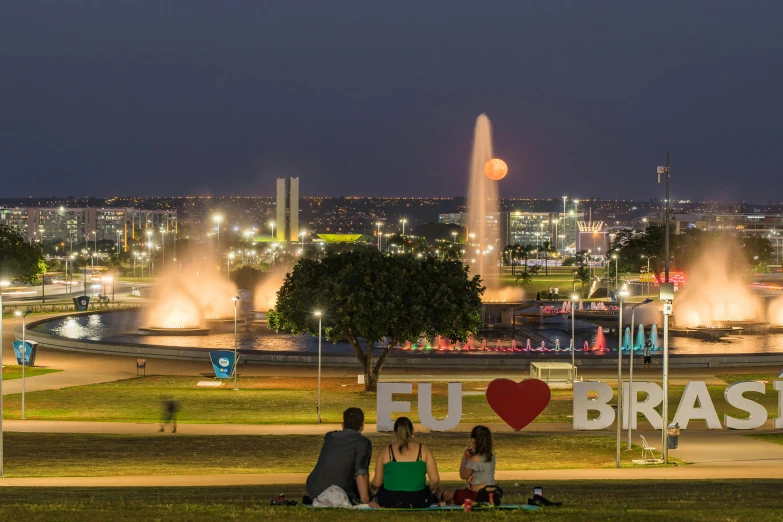 a group of people sitting on top of a lush green field, brutalism, city lights on the horizon, fountains, enso, on a hot australian day