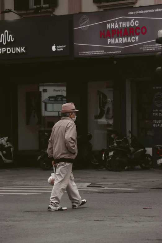 a man walking across a street in front of a pharmacy, by Niko Henrichon, trending on unsplash, an oldman, grey clothes, caracter with brown hat, low quality photo