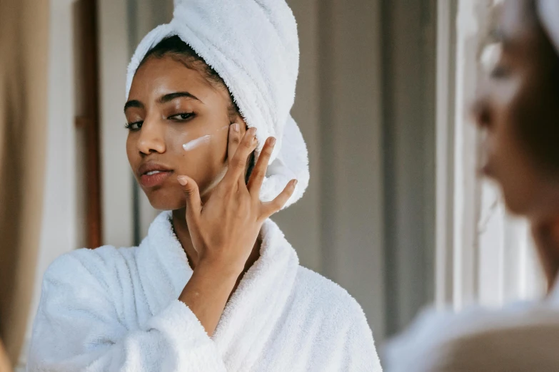 a woman putting cream on her face in front of a mirror, trending on pexels, renaissance, wearing long white robe, manuka, indian girl with brown skin, wearing a towel