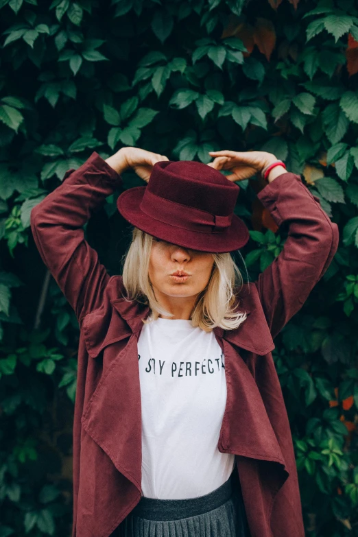 a woman standing in front of a bush with her hands on her head, by Julia Pishtar, pexels contest winner, tipping his fedora, graphic tees, maroon, a blond