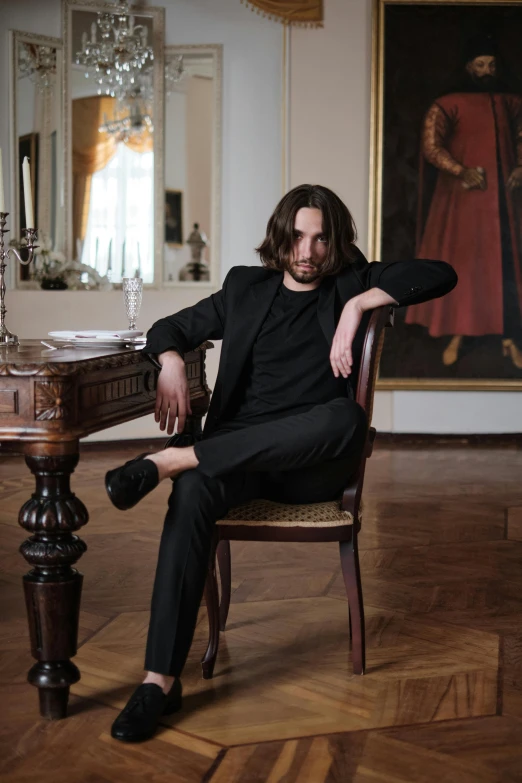 a man sitting in a chair in front of a painting, an album cover, inspired by Ramon Pichot, pexels contest winner, baroque, mid long hair, wearing black clothes, andrea savchenko, christian bale