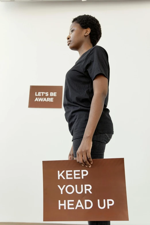 a woman holding a sign that says keep your head up, an album cover, by Lily Delissa Joseph, standing posture, 2010s, ( ( brown skin ) ), emergency