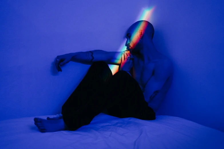 a man sitting on top of a bed under a rainbow light, an album cover, inspired by Elsa Bleda, pexels contest winner, holography, hairless, profile image, red and blue black light, abstract human body