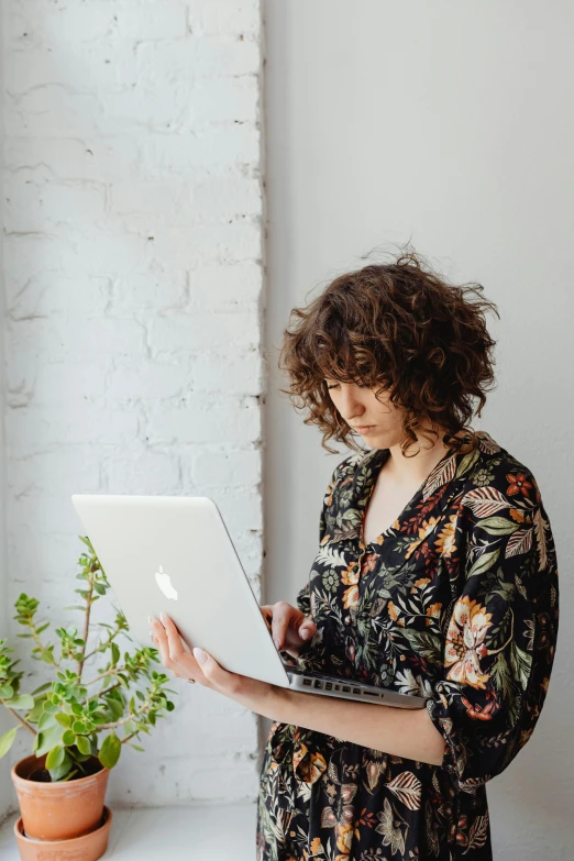 a woman standing in front of a window holding a laptop, pexels contest winner, renaissance, curly and short top hair, profile image, standing on a desk, floral patterned skin