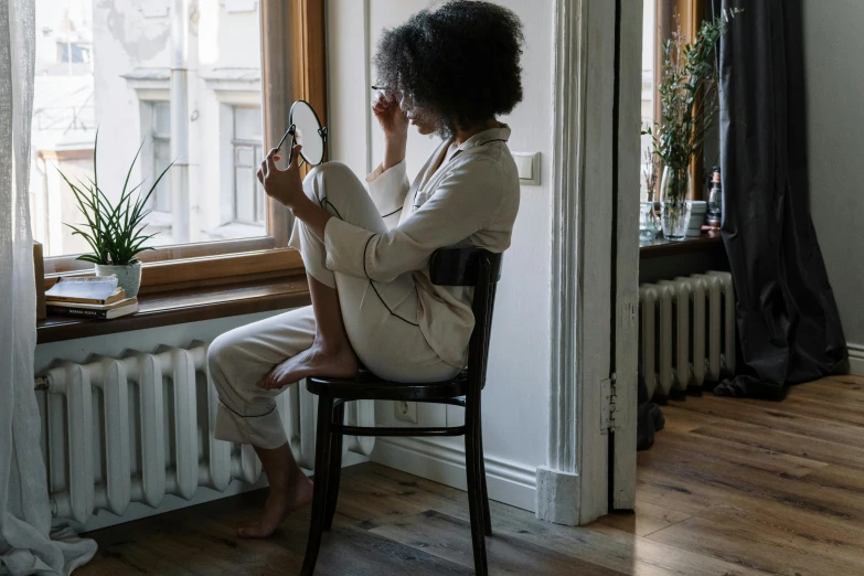 a woman sitting in a chair looking at her reflection in a mirror, by Nina Hamnett, pexels contest winner, renaissance, wearing pajamas, with brown skin, lonely and sad, near the window