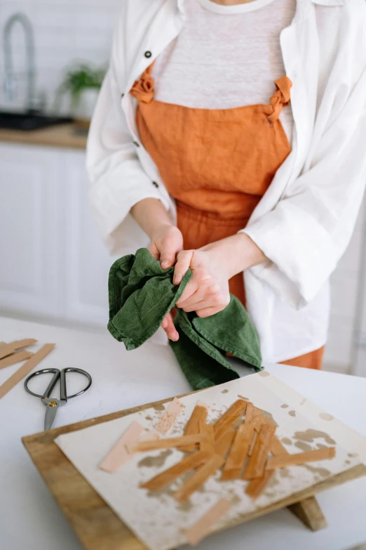 a woman standing in a kitchen next to a cutting board, inspired by Maki Haku, trending on pexels, process art, folds of fabric, copper oxide and rust materials, scissors in hand, dressed as a pastry chef