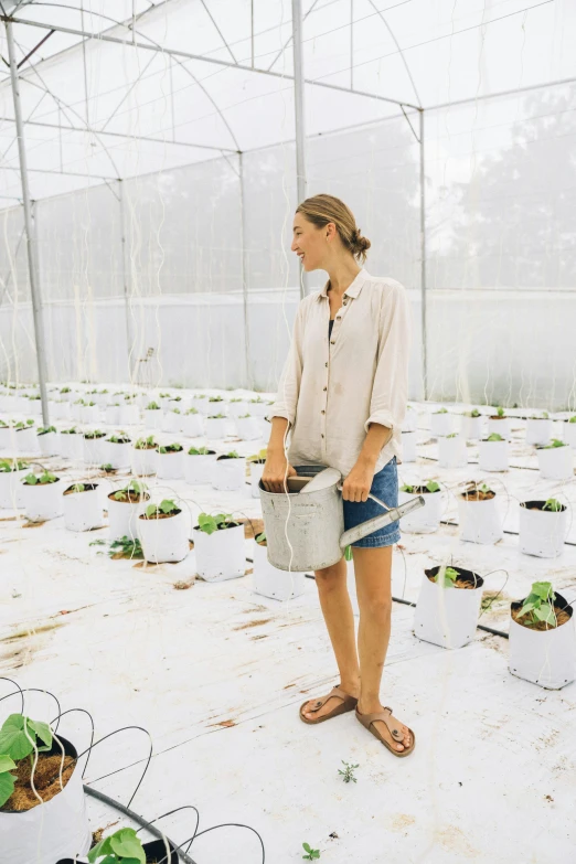 a woman standing in a greenhouse holding a watering can, unsplash, happening, wearing a linen shirt, hydroponic farms, australian, bag over the waist