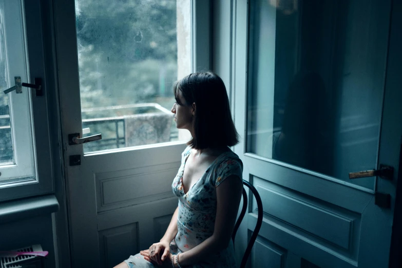 a woman sitting in a chair looking out a window, a picture, inspired by Elsa Bleda, unsplash contest winner, realism, wearing a plastic blue dress, vietnamese woman, medium format. soft light, anna nikonova aka newmilky