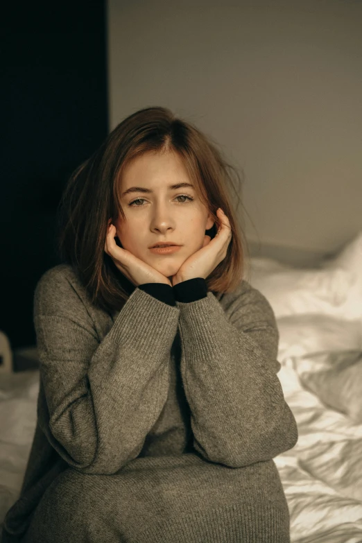 a woman sitting on a bed with her hands on her face, trending on pexels, wearing a dark sweater, sad face, girl with brown hair, grey