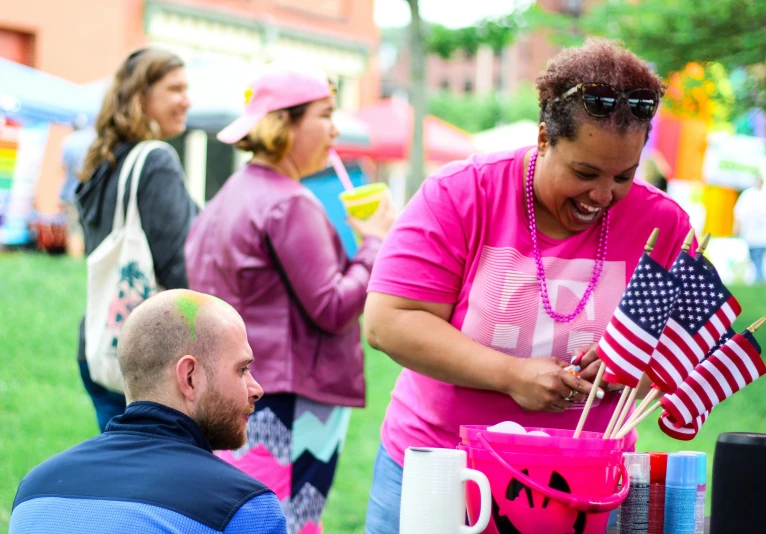 a group of people standing around a table with american flags, a photo, fluorescent pink face paint, people on a picnic, vendors, pink