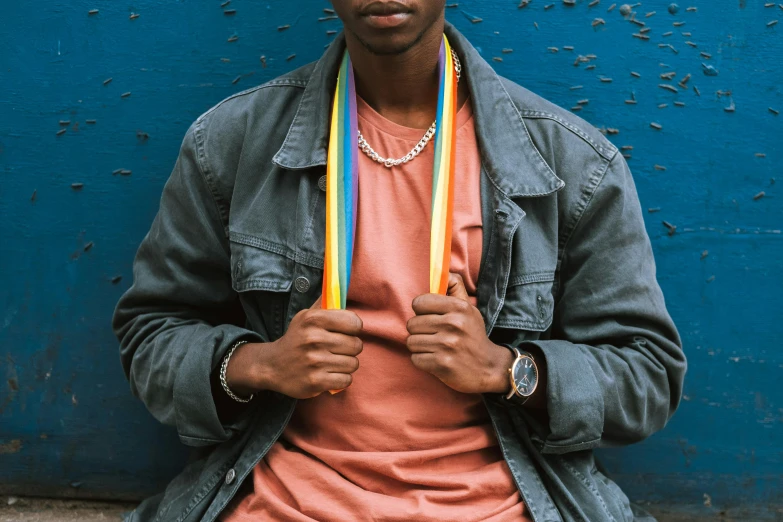 a man standing in front of a blue wall, an album cover, trending on pexels, black arts movement, rainbow stripe backdrop, bracelets and necklaces, scarf made from spaghetti, male teenager