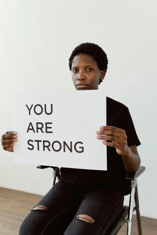 a woman sitting in a chair holding a sign that says you are strong, by Dulah Marie Evans, dada, white ribbon, david uzochukwu, high quality photo, lgbt