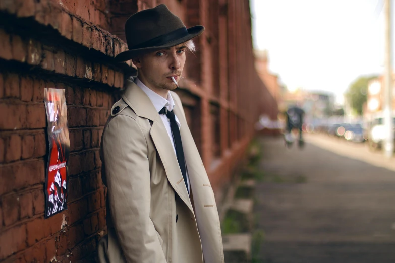 a man leaning against a brick wall with a cigarette in his mouth, inspired by John Watson Gordon, wearing a long beige trench coat, lachlan bailey, pork pie hat, model photograph