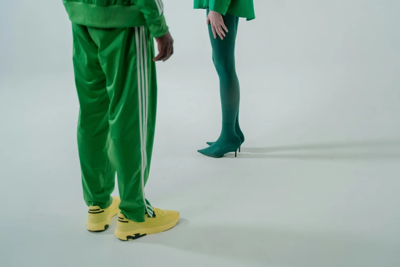 a woman standing next to a man in a green suit, inspired by Elsa Bleda, antipodeans, futuristic balenciaga sneakers, chappie in an adidas track suit, showstudio, with yellow cloths